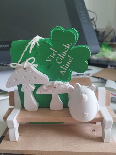 Viel Glück Geschenk Holzbank mit weißen Stützen, zwei unterschiedlichen Fliegenpilzen, Bärchen und einer Karte in Kleeblattform mit Viel Glück