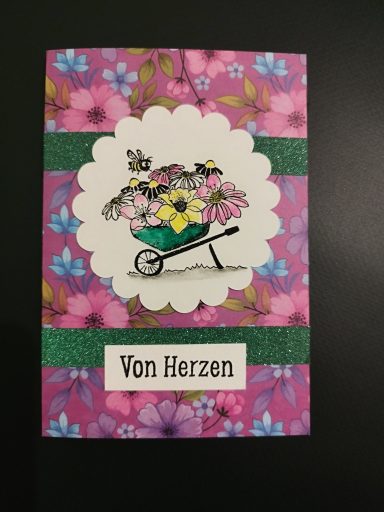 Karte "Von Herzen" pinke Blumenkarte, mit Schubkarre voller Blumen und Bienchen, abgesetzt mit glitzernden grünen Bändern.